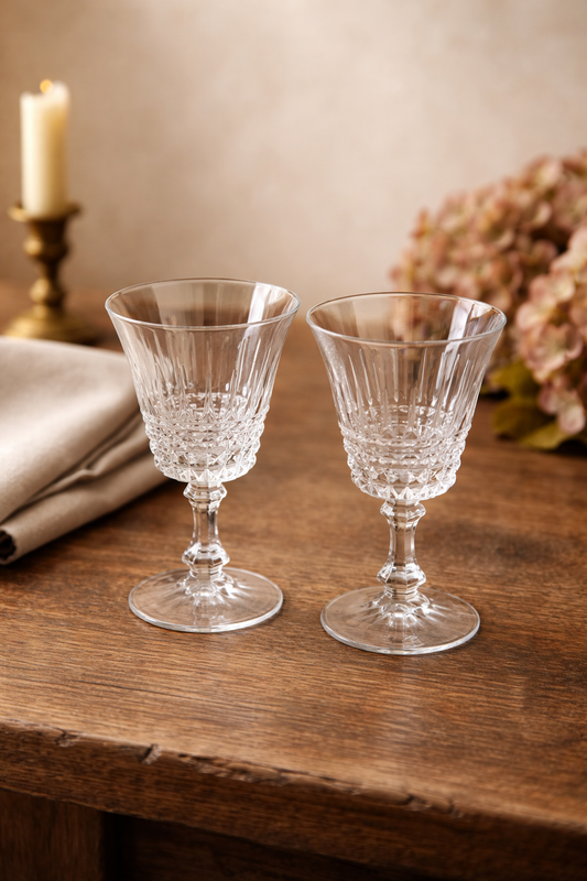 Two crystal glasses on a wooden table with a candle and flowers in the background.